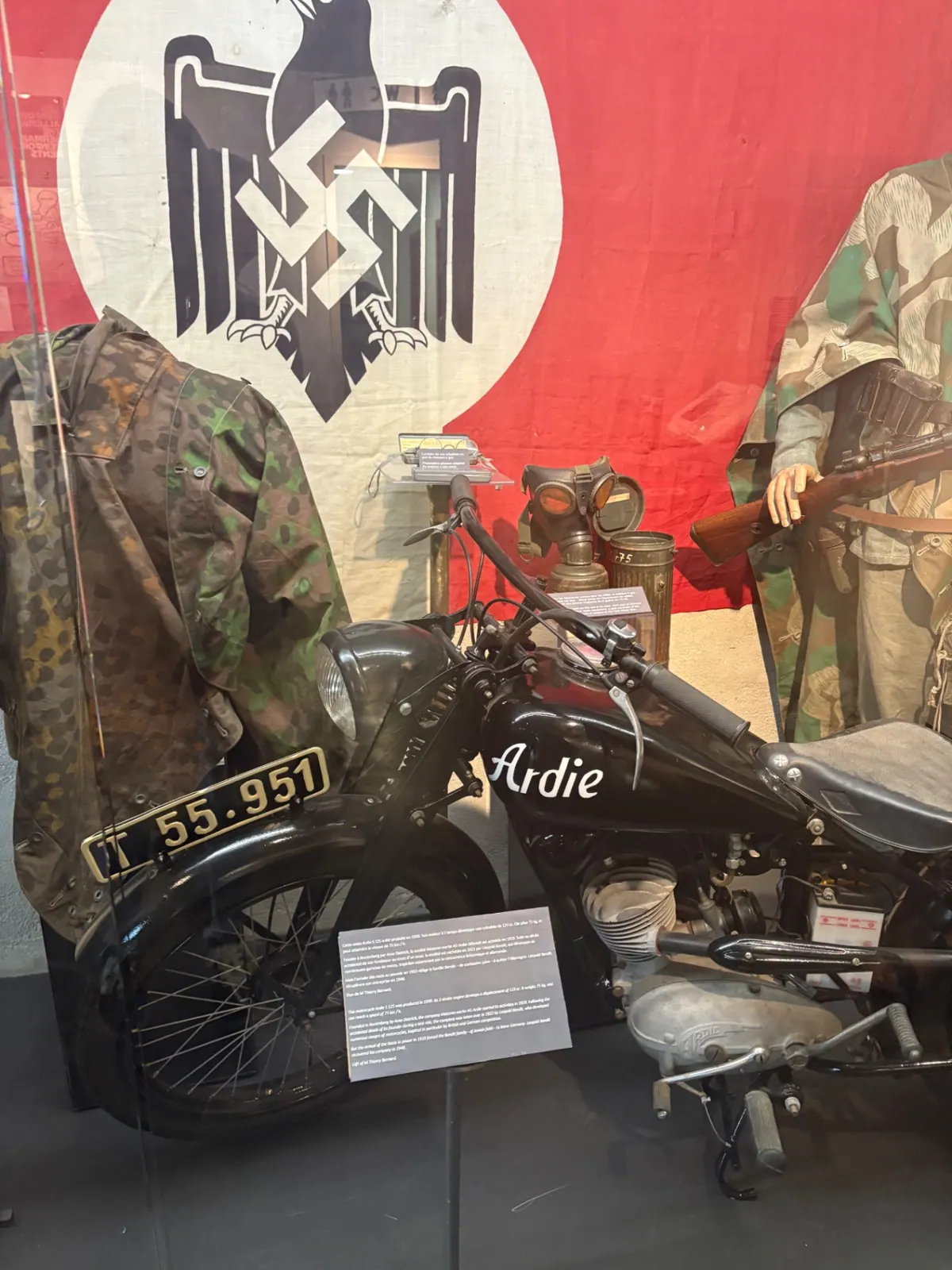 Museum case with a German Ardie motorcycle, camouflage smocks and a gas mask in front of a Reich flag