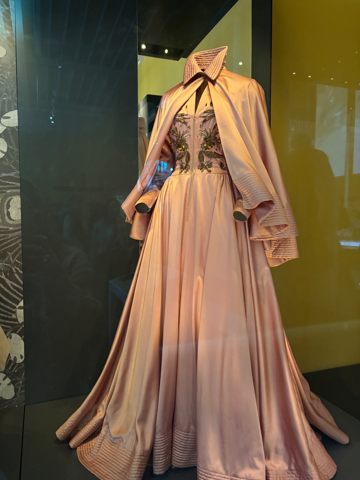 A rose-gold embroidered gown and cape displayed behind museum glass