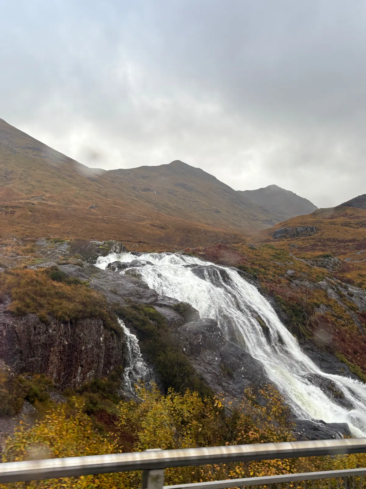 A wide waterfall pouring over dark rock in the rust-colored hills of Glencoe