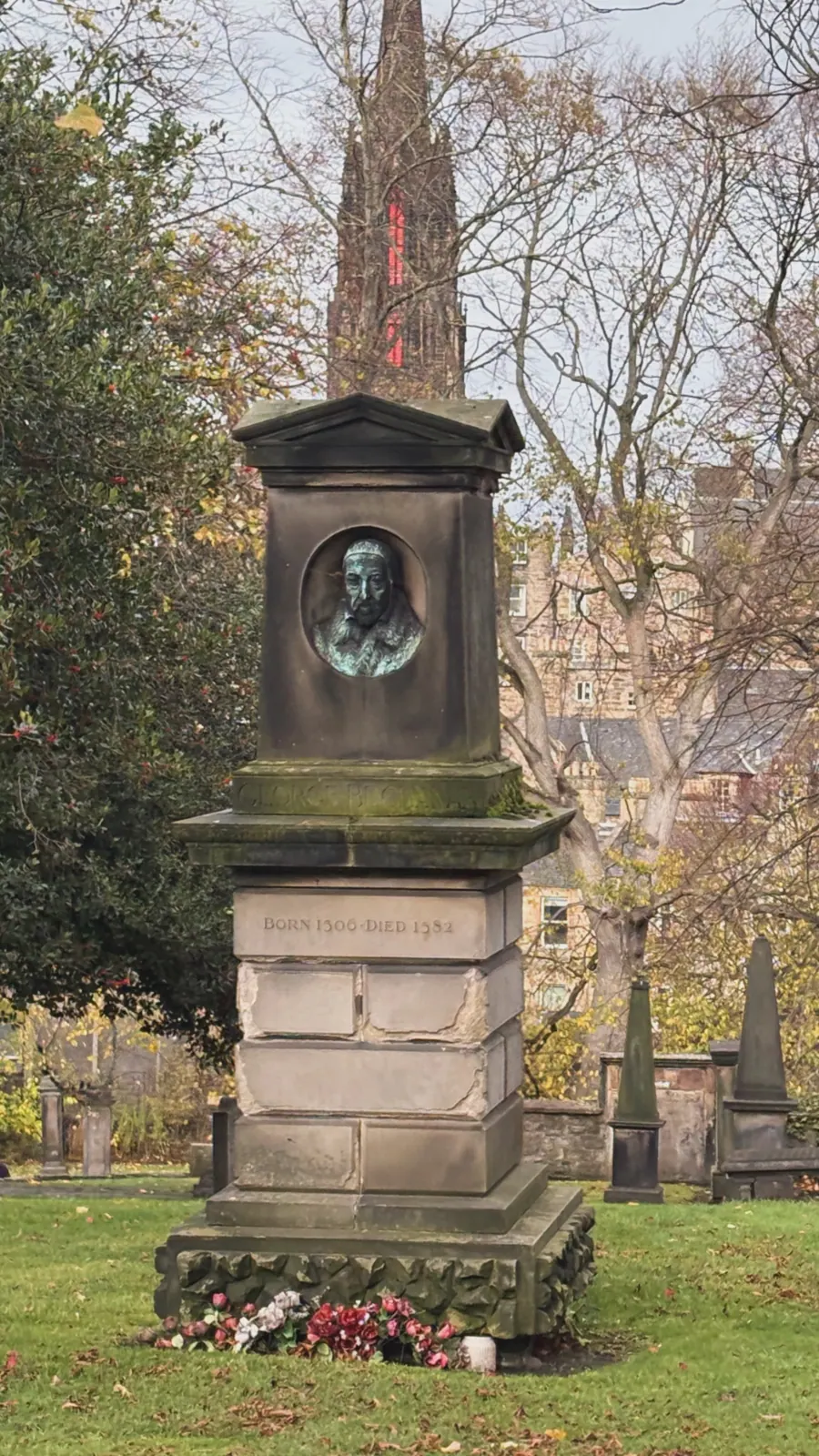 A weathered stone monument with a bronze portrait bust, flowers laid at its base